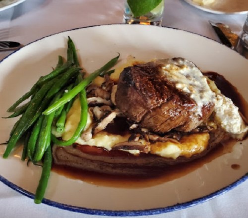 A plated meal featuring a beef filet on mashed potatoes, topped with cheese, alongside green beans and a rich sauce.