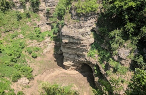 A rocky cliff surrounded by lush greenery, with a cave entrance visible at the base and a dry area below.