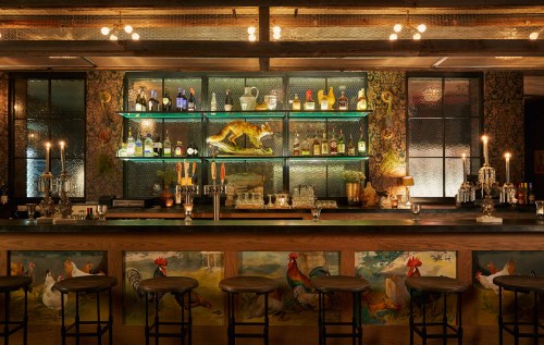 A stylish bar with a wooden counter, colorful decor, shelves of bottles, and candlelight ambiance.