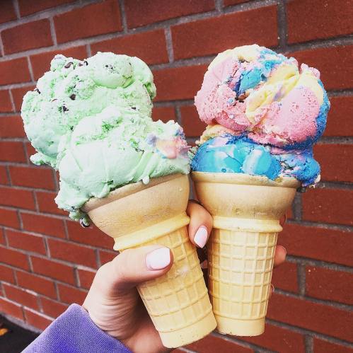 A hand holding two ice cream cones, one with green and pink scoops, the other with blue and yellow scoops, against a brick wall.