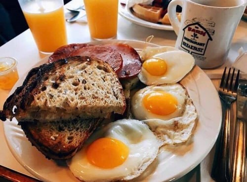 A plate of breakfast featuring toast, fried eggs, and slices of ham, with orange juice in the background.