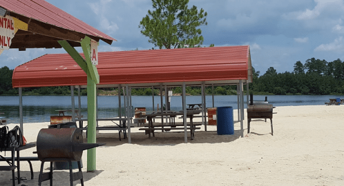 Sheltered picnic area by a sandy beach, with grills and tables overlooking a calm lake under a cloudy sky.