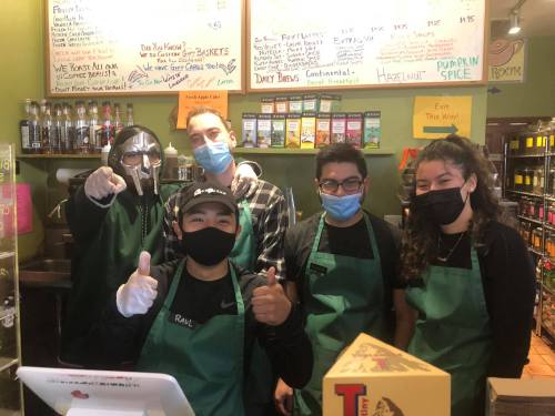 A group of five masked employees in green aprons pose together at a counter, smiling and giving thumbs up.