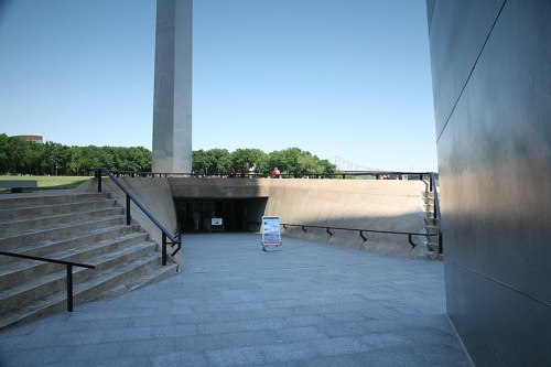 A view of a modern building entrance with stairs, leading to a lower level, and a tall monument in the background.