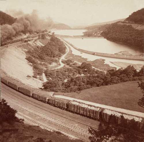 A vintage landscape showing a river, train tracks, and a train near a mountainous area with smoke in the background.