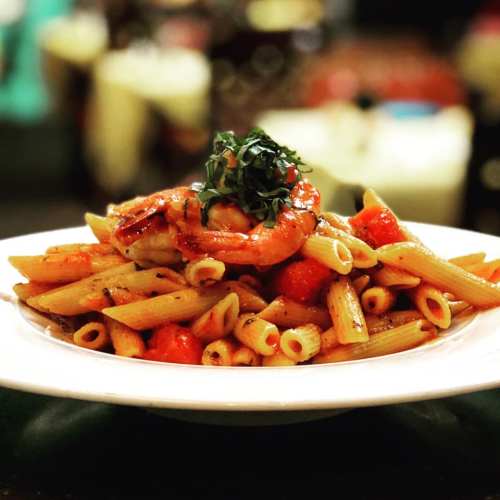 A plate of penne pasta topped with shrimp, cherry tomatoes, and fresh basil, served in a restaurant setting.