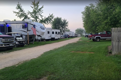 A gravel road lined with RVs and trucks, surrounded by green grass and trees in a camping area.