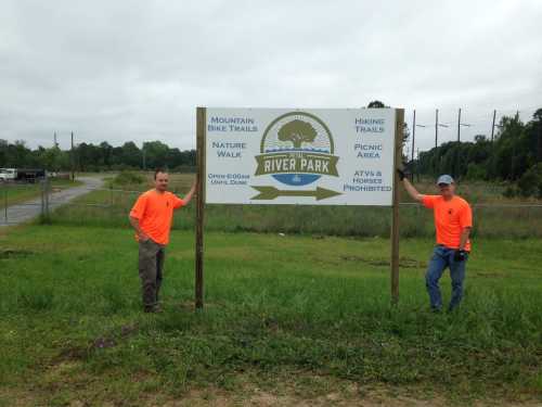 Two men in orange shirts stand beside a sign for River Park, listing activities like hiking and biking trails.