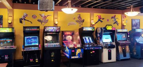 A row of vintage arcade machines against a yellow wall featuring superhero artwork.