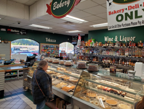 A woman stands in a bakery shop, looking at a display of pastries and desserts, with shelves of liquor and tobacco nearby.