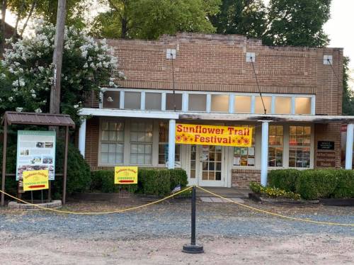 A brick building with a banner reading "Sunflower Trail Festival" and signs for a local museum, surrounded by greenery.