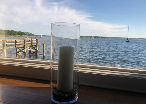 A candle in a glass holder sits on a windowsill, overlooking a calm bay with boats and a distant shoreline.