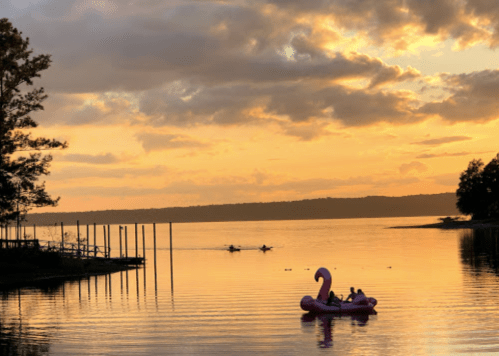 A serene sunset over a lake, with two people on a pink float and kayakers in the distance.