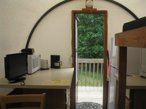 Interior of a cozy cabin with a kitchenette, desk, and door leading to a wooden deck surrounded by greenery.