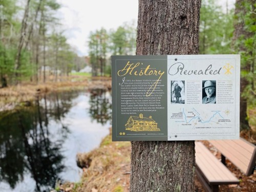 A sign on a tree by a calm pond, detailing local history, surrounded by trees and a wooden bench nearby.