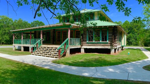 A large, two-story log cabin with a green roof, surrounded by trees and a paved walkway.
