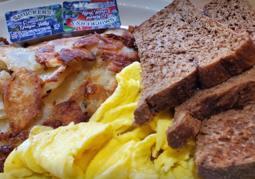 A plate with scrambled eggs, crispy hash browns, and slices of brown bread, accompanied by jelly packets.