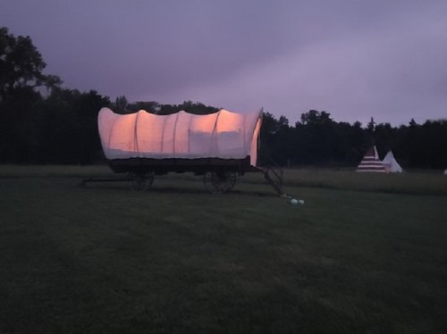A lit covered wagon on wheels in a grassy field, with teepees visible in the background under a twilight sky.