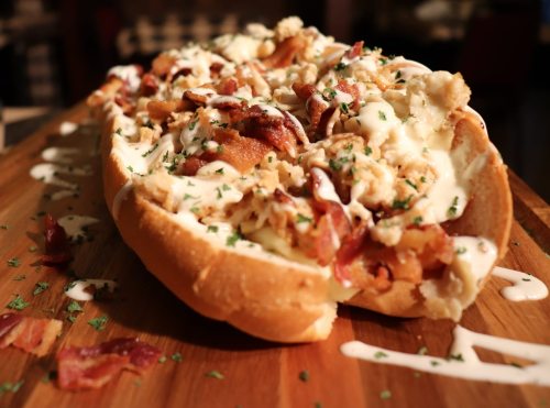 A loaded sandwich on a wooden board, topped with bacon, chicken, and drizzled with creamy sauce and herbs.