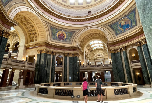 Two people stand in a grand, ornate hall with high ceilings and intricate architectural details.