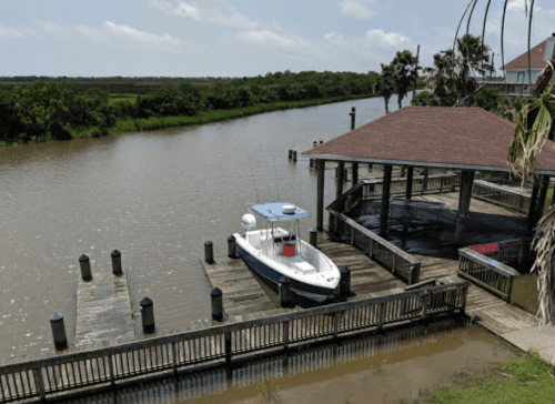 A boat docked at a wooden pier beside a calm river, surrounded by lush greenery and a clear blue sky.