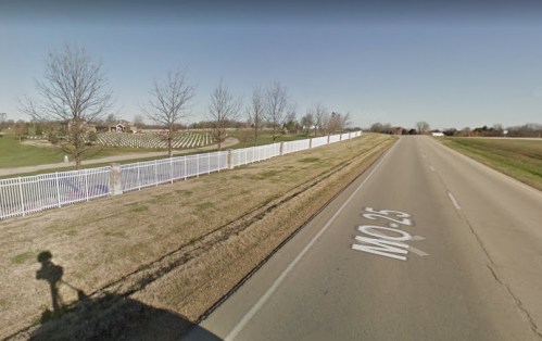 A wide road with a white fence alongside, leading to a grassy area with trees and a distant cemetery.