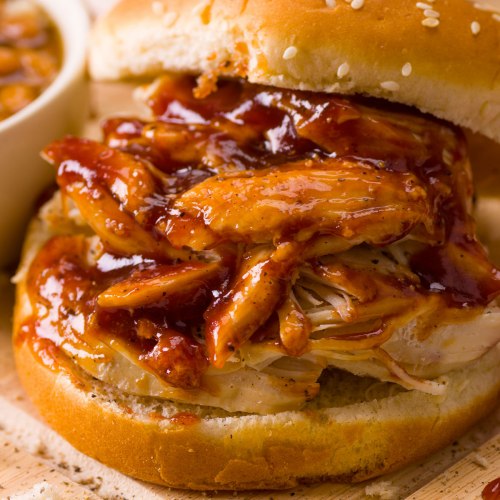 A close-up of a sesame seed bun filled with shredded chicken and barbecue sauce, served with a side of dipping sauce.