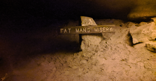 A wooden sign reading "Fat Man's Misery" placed on rocky ground in a dimly lit cave.