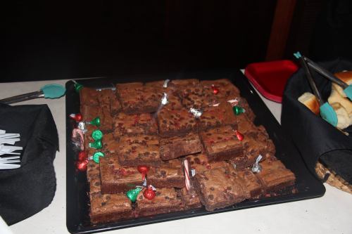 A tray of chocolate brownies decorated with red and green candies, placed on a table with other snacks.