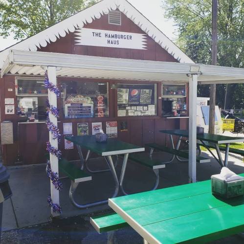 A small, rustic hamburger stand with green picnic tables and a sign reading "The Hamburger Haus."