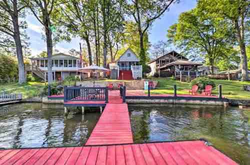 A scenic lakeside view featuring a red dock, green trees, and charming houses with outdoor seating.