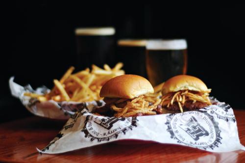 Two sliders topped with crispy onions on a paper-lined plate, with fries and two glasses of beer in the background.