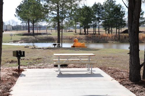 A picnic table near a pond with a large yellow duck and trees in the background on a sunny day.
