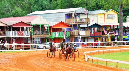 A colorful horse racing scene with jockeys on a dirt track, surrounded by vibrant buildings and spectators.