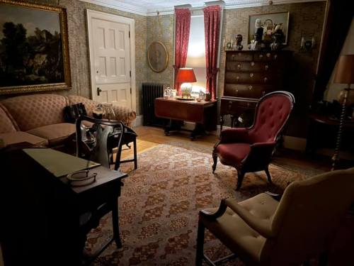 Cozy vintage living room with patterned wallpaper, a red armchair, a wooden desk, and warm lighting.