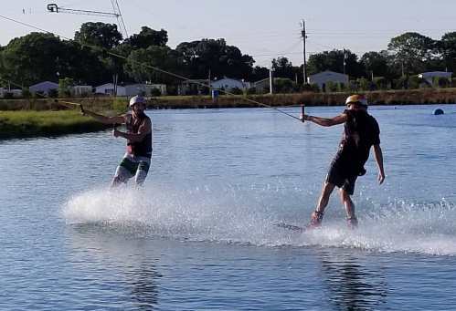 Two people wakeboarding on a calm lake, creating splashes as they glide across the water under a clear sky.