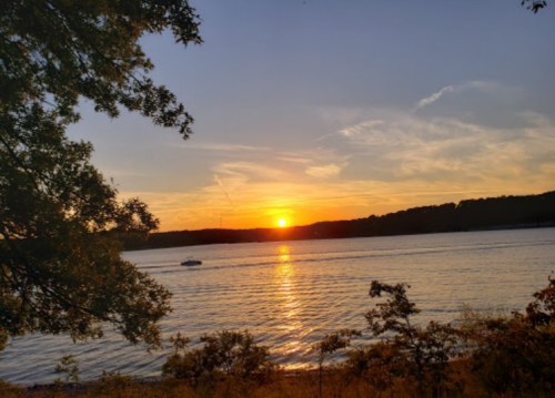 A serene sunset over a calm lake, with a boat gliding across the water and trees framing the scene.