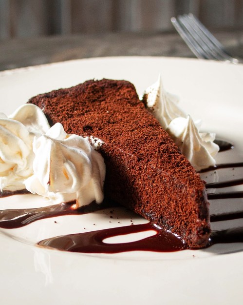 A slice of rich chocolate cake topped with whipped cream and drizzled with chocolate sauce on a white plate.