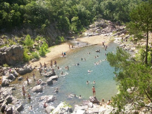 A scenic river with people swimming and relaxing on the rocky shore, surrounded by lush greenery and trees.