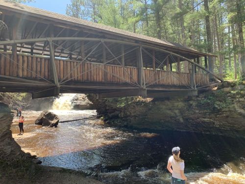 A wooden bridge spans a river with people standing nearby, surrounded by trees and a waterfall in the background.