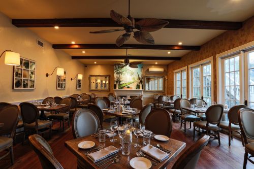 Cozy restaurant interior with wooden tables, elegant chairs, and large windows, featuring warm lighting and tropical decor.