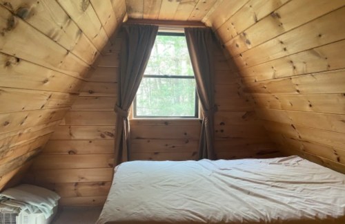 Cozy attic bedroom with wooden walls, a window, and a bed without bedding. Natural light filters in through the curtains.
