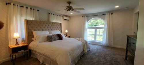 Bright, cozy bedroom with a large bed, nightstands, and a window featuring sheer curtains. Soft carpet and ceiling fan.