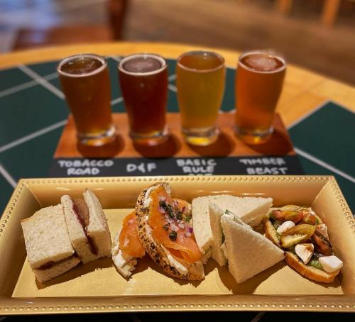 A platter of assorted sandwiches and a small dish, accompanied by four glasses of craft beer on a wooden flight board.