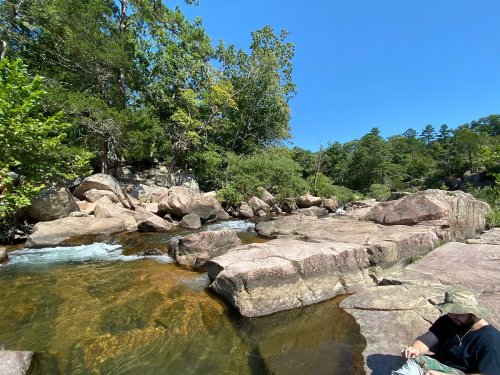A serene river scene with clear water flowing over rocks, surrounded by lush greenery and a blue sky.