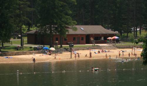 A sandy beach by a lake with people swimming, sunbathing, and a building in the background surrounded by trees.