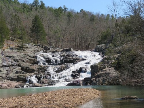 A serene waterfall cascades over rocky terrain, surrounded by trees and a clear blue sky.