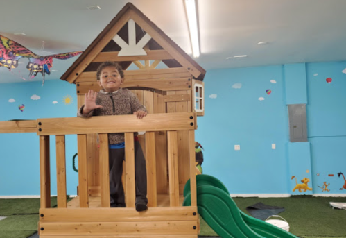 A child smiles and waves from a wooden playhouse with a slide in a colorful indoor play area.
