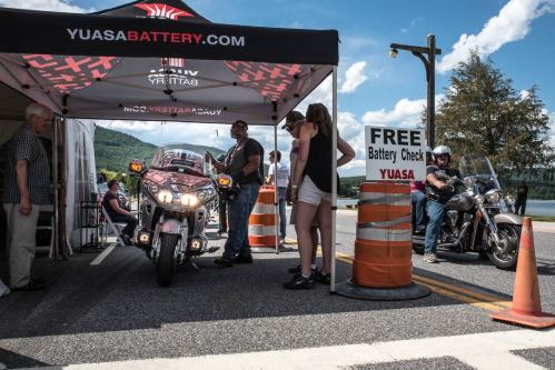 A motorcycle under a tent at a battery check event, with people waiting and a sign for free battery checks nearby.