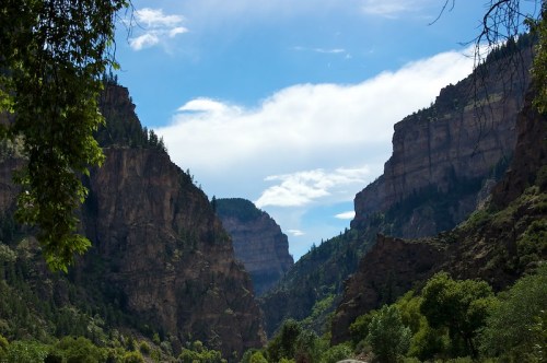 A scenic view of a canyon with steep cliffs, lush greenery, and a bright blue sky with scattered clouds.